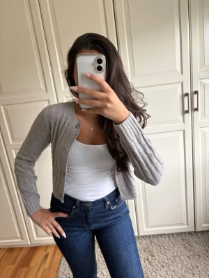 Grå ribbstickad kofta - Snygg grå ribbstickad kofta med rund halsringning och knappar framtill. Kortare modell som passar perfekt till jeans eller kjol. Tillverkad i mjukt material som känns skönt mot huden. Enkel och stilren design som är lätt att matcha.