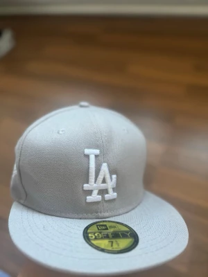  LA-keps från New Era - Snygg ljusgrå keps från New Era med klassisk LA-logga broderad i vitt framtill. Modellen är 59FIFTY med rak skärm och strukturerad passform. Perfekt för dig som gillar streetwear och sportig stil.
