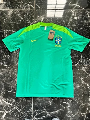 Grön Brasilien Nike träningströja - Snygg grön träningströja från Nike med Brasilien CBF-logga och gula detaljer. Tröjan har korta ärmar, rund hals och är tillverkad i ett lätt, ventilerande material som passar perfekt till fotbollsträning eller gymmet.