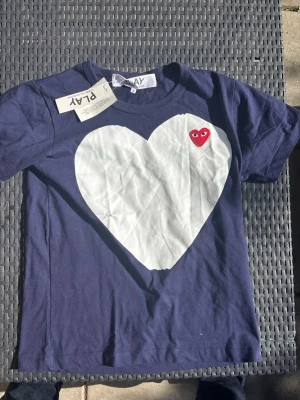 Mörkblå t-shirt med hjärta - Snygg mörkblå t-shirt med ett stort vitt hjärta på bröstet och en liten röd hjärtdetalj med ögon. Klassisk rund hals och korta ärmar. Tillverkad i mjuk bomull, perfekt för en avslappnad stil.