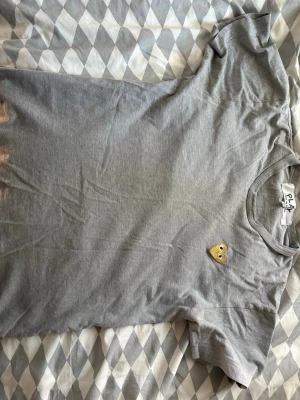Grå t-shirt med gul hjärtdetalj - Säljer en grå t-shirt med korta ärmar och rund hals. På bröstet sitter en liten gul hjärtdetalj som sticker ut och ger tröjan en unik touch. Perfekt till jeans eller shorts för en enkel och clean look.