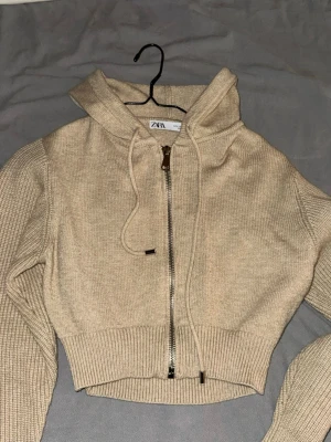 Beige stickad kofta från Zara - Säljer en beige stickad kofta från Zara med dragkedja framtill och ribbade muddar. Tröjan har långa ärmar, huva med dragsko och croppad passform. Perfekt för dig som gillar en avslappnad men ändå trendig stil.