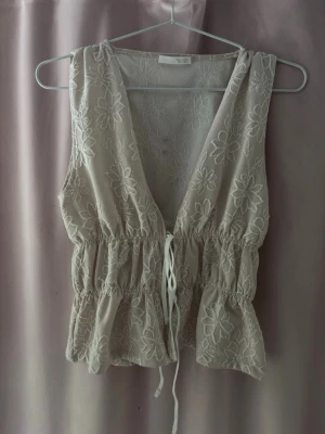 Beige broderad blus från Zara - Supersöt beige blus från Zara med broderade blommor och knyt i midjan. Ärmlös modell med v-ringning och volangkant nedtill. Perfekt för dig som gillar romantiska detaljer och vill ha en luftig topp till sommaren.