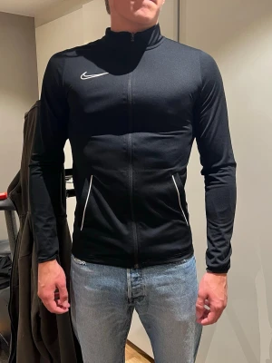 Svart träningströja från Nike - Snygg svart träningströja från Nike med hel dragkedja framtill och vita detaljer vid fickorna samt Nike-logga på bröstet. Jackan har hög krage och är tillverkad i ett stretchigt funktionsmaterial som passar perfekt till träning. Tröjan har en trasig sömn/ resår vid höger arm.