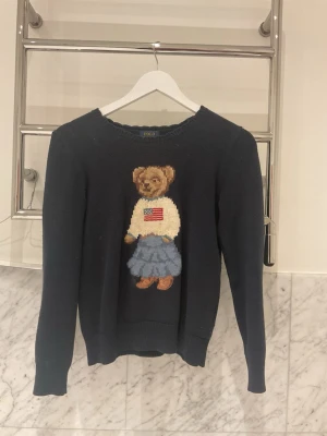 Mörkblå stickad tröja med björn - Mysig mörkblå stickad tröja med en stor broderad björn på framsidan. Björnen har en vit tröja med amerikansk flagga och blå kjol. Tröjan har rund hals och långa ärmar, perfekt för dig som gillar klassiska och lekfulla detaljer.