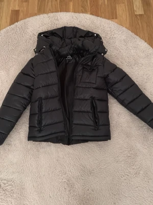 Svart pufferjacka med huva - Snygg svart pufferjacka med huva och dragkedja framtill. Jackan har två framfickor med dragkedja och quiltad design. Perfekt för kalla dagar och har en modern, sportig look. Materialet är glansigt och ger en trendig vibe.