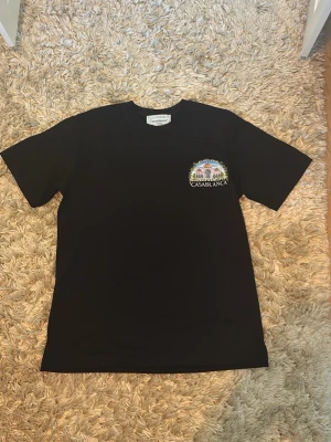 Svart Casablanca t-shirt med tryck - Säljer en svart t-shirt från Casablanca med färgglatt tryck av byggnad och palmer på ryggen samt mindre tryck på bröstet. Klassisk passform och rund hals. Perfekt för dig som vill ha en stilren men ändå unik look.