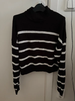 Svartvit randig polotröja - Säljer en svart stickad polotröja med vita horisontella ränder. Tröjan har lång ärm och hög krage, vilket ger en klassisk och stilren look. Perfekt för dig som gillar enkel men snygg design. Materialet känns mjukt och bekvämt.