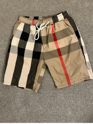 Rutiga shorts med snörning - Snygga shorts med stort rutmönster i beige, svart, vitt och rött. De har elastisk midja med vit snörning och sidofickor. Perfekta för sommaren och ger en trendig look.