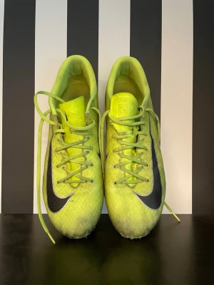  Nike Air Zoom fotbollsskor med dobbar - Säljer ett par Nike fotbollsskor med svart swoosh på sidan. Skorna har snörning och är tillverkade i ett lätt syntetmaterial för bra bollkontroll. Dobbarna är anpassade för spel på gräs. Perfekta för dig som vill sticka ut på planen.