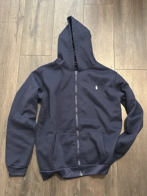 Mörkblå hoodie med dragkedja - Snygg mörkblå hoodie med dragkedja framtill och två stora fickor. Klassisk design med liten vit logga på bröstet och ribbade muddar vid ärmar och nederkant. Tillverkad i mjukt bomullsmaterial och har en stor huva för extra komfort.