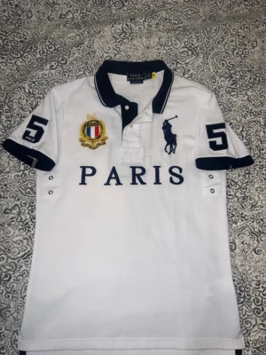 Vit pikétröja Polo Ralph Lauren Paris - Vit pikétröja från Polo Ralph Lauren med svart krage och ärmslut. Stora tryck och broderier framtill, bland annat PARIS och en guldfärgad patch. Siffran 5 på båda ärmar och ryggen. Klassisk pikéknäppning och kort ärm. Perfekt för en sportig och stilren look.