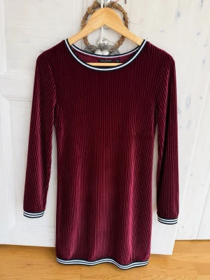 Vinröd sammetklänning Soul River - Större i storleken så passar även s. Vinröd kortklänning i mjuk sammet från Soul River. Klänningen har långärmad design, ribbad struktur och sportiga kontrastband i svart och vitt vid halsringning och ärmslut. Perfekt för dig som gillar en stilren men ändå trendig look.