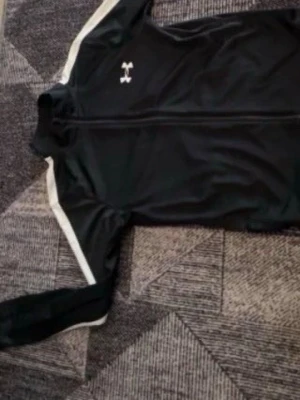 Svart träningsjacka från Under Armour - Snygg svart träningsjacka från Under Armour med hel dragkedja framtill och vita detaljer längs ärmarna. Jackan har en hög krage och är tillverkad i ett lätt, stretchigt material som passar perfekt till träning eller uppvärmning.