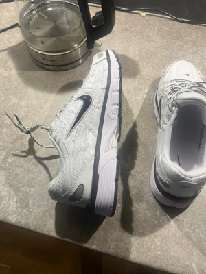 Nike vita sneakers med svart swoosh - Vita sneakers från Nike med svart swoosh på sidan. Skorna har ovandel i mesh och syntet, vita snören och en kraftig sula i vitt och svart. Sportig design med detaljerade sömmar och ventilerande material.