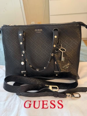 Svart handväska från Guess - Snygg svart handväska från Guess med präglat logomönster och silverfärgade detaljer. Väskan har både handtag och avtagbar axelrem samt dekorativ nyckelring. Perfekt för dig som vill ha en stilren och trendig accessoar. Dustbag medföljer. 