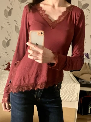 Vinröd långärmad topp med spets - Vinröd långärmad topp med v-ringning och spetsdetaljer vid hals, ärmslut och nederkant. Modellen är figurnära med bekvämt material 