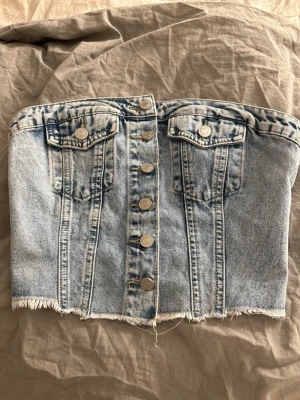 Ljusblå denim korsett tubtopp - Trendig ljusblå denim korsett tubtopp med knappar framtill, två bröstfickor och rå fransad kant nedtill. Baksidan har smockdetalj för extra stretch och passform. Perfekt för en edgy och cool look.