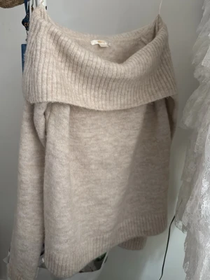 Beige off shoulder stickad tröja - Mysig beige stickad tröja med bred off shoulder-krage och ribbade detaljer vid hals, ärmslut och nederkant. Tröjan har en avslappnad passform och är perfekt för lager på lager. Materialet ser mjukt och fluffigt ut, vilket ger en skön känsla.
