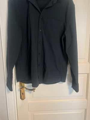 Svart långärmad skjorta med ficka - Stilren mörkgrön skjorta med lång ärm och klassisk krage. Skjortan har en bröstficka och knäppning framtill. Perfekt till dig som gillar en enkel och clean look.