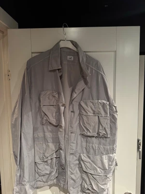 Ljusgrå overshirt från C.P. Company - Säljer en ljusgrå overshirt från C.P. Company med fyra stora fickor framtill och klassisk krage. Overshirten har knappstängning och är tillverkad i ett tunt, lätt material som passar perfekt för lager-på-lager. Snygg och funktionell design.