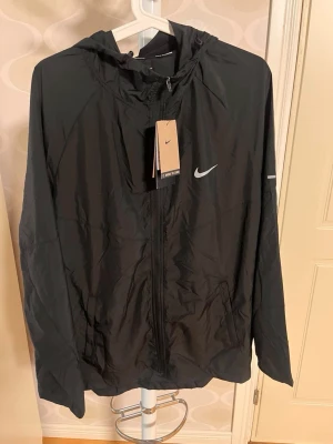 Svart vindjacka från Nike med huva - Snygg svart vindjacka från Nike med dragkedja framtill, huva och två sidofickor. Jackan har en diskret Nike-logga på bröstet och är tillverkad i lätt, vindtätt material. Perfekt för blåsiga dagar och enkel att matcha med olika outfits.