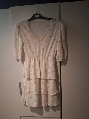 Vit blommig volangklänning - Söt vit kortklänning med små blå blommor, v-ringning och trekvartsärm. Klänningen har flera volanger i kjolen och är gjord i ett lätt, luftigt material. Perfekt för varma dagar och somriga tillfällen. Använd 1 gång 