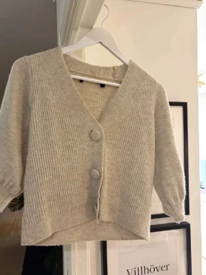 Beige ribbstickad kofta med knappar - Superfin beige kofta med ribbstickat mönster och två stora knappar framtill. Koftan har V-ringning och lite kortare, boxig passform med halvlånga ärmar. Perfekt att slänga över en topp för en chill look.