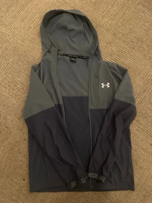 Under Armour grå och mörkblå vindjacka (OBS dragkedja bör fixas) - Snygg vindjacka från Under Armour i två nyanser av grått och mörkblått. Jackan har huva, dragkedja framtill och logga på bröstet. Tillverkad i lätt och smidigt material, perfekt för blåsiga dagar. Elastiska muddar med Under Armour-tryck nertill.