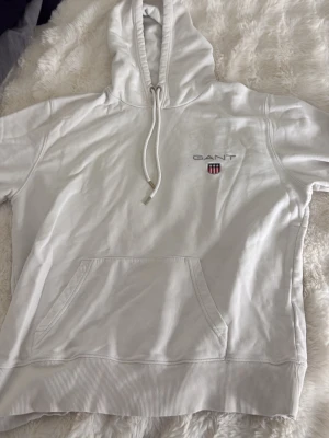 Vit hoodie från GANT med logga - Snygg vit hoodie från GANT med broderad logga och sköld på bröstet. Klassisk känguruficka framtill och justerbar dragsko i huvan. Tillverkad i mjukt bomullsmaterial som känns skönt mot huden. Perfekt för en avslappnad och stilren look.