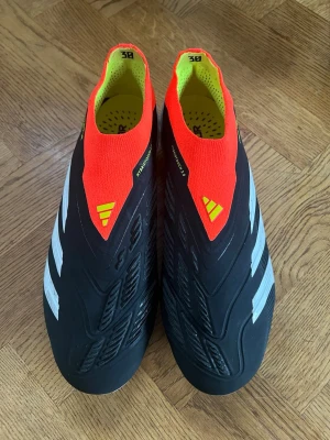 Adidas Predator fotbollsskor svart/röd - Säljer ett par Adidas Predator fotbollsskor med svart bas, vita detaljer och röda samt gula inslag. Skorna har en strumpliknande passform utan snörning och är tillverkade i syntetmaterial med Hybridtouch 2.0-teknologi för bättre bollkontroll. Använda en gång, nyskick! 