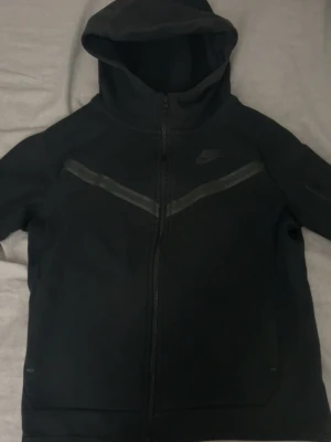 Svart Nike Tech Fleece hoodie - Svart Nike Tech Fleece hoodie med dragkedja, huva och två fickor fram. Snygga reflexdetaljer över bröstet och en extra ficka med dragkedja på ärmen. Perfekt för dig som gillar sportig och clean stil.