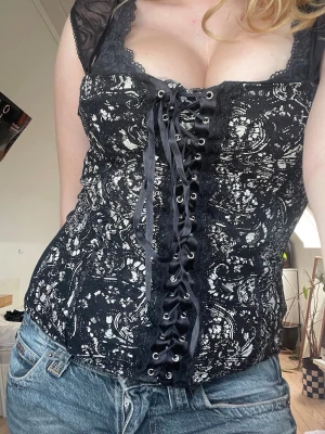 Svart och vit korsetttröja med snörning - Trendig svart korsetttröja med vitt mönster, spetsdetaljer och snörning framtill. Toppen har meshpartier vid axlarna och en figurnära, slim passform. Perfekt för att skapa en cool och edgy stil. Tröjan är mjuk och är inte var korsett material som man kan se på baksidan💕