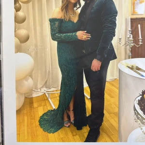 Dressroom bal - Elegant långklänning i mörkgrön spets med offshoulder och lång ärm. Klänningen har en hög slits framtill och figurnära passform. Perfekt för speciella tillfällen och ger en lyxig känsla.