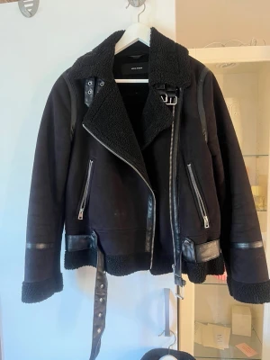 Svart skinnjacka från Vero Moda - Cool svart skinnjacka från Vero Moda med bred krage i fuskpäls, detaljerade dragkedjor och bälte i midjan. Jackan har en mix av skinn och teddy-material, vilket ger en trendig look. Perfekt för dig som vill ha en edgy stil.