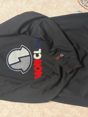 Svart Moncler sweatshirt med logga - Svart sweatshirt från Moncler med stor broderad logga och text på bröstet i rött, vitt och mörkgrönt. Tröjan har rund hals, långa ärmar och ribbade muddar. Extra Moncler-detalj på ena ärmen. Perfekt för en trendig streetwear-look.