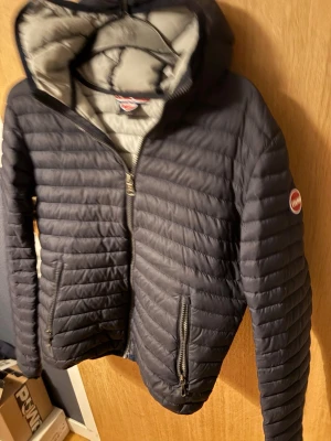 Colmar jacka  - Snygg mörkblå dunjacka från Colmar med huva och dragkedja framtill. Jackan har två fickor med dragkedja och ett stort Colmar-märke på ärmen. Insidan är ljusgrå och jackan har en quiltad design som ger en sportig look.
