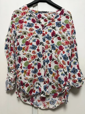 Blommig och fruktmönstrad blus från Cecil - Färgglad blus med lång ärm från Cecil, täckt av blommor och frukter som kiwi, granatäpple och vindruvor på vit botten. Blusen har knappar framtill och dragsko vid nederkanten. Perfekt för dig som gillar unika och livliga mönster.