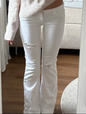 Vita bootcut jeans med slitningar - Säljer ett par vita bootcut jeans med slitningar vid knäna och snygga broderade bakfickor. Jeansen har hög midja och klassisk femficksdesign. Perfekta för dig som vill ha en trendig och avslappnad look. (Lånar bild av personen jag köpte av) säljer då dem ba lite lite för tajta vid midjan! 