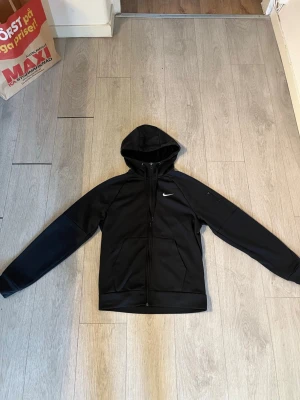  Nike hoodie  - Snygg svart hoodie från Nike med dragkedja framtill och klassisk vit Nike-logga på bröstet. Tröjan har huva och två fickor på sidorna. Tillverkad i ett mjukt och bekvämt material som passar perfekt till en sportig stil.