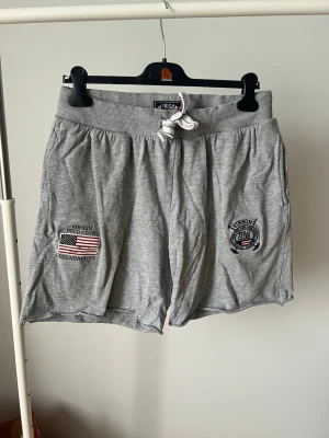 Grå shorts med snörning från Vinson - Grå mjuka shorts från Vinson med snörning i midjan och tryck med amerikansk flagga och text på framsidan. Shortsen har elastisk midja, fickor fram och en bakficka. Perfekta för chill dagar eller träning.