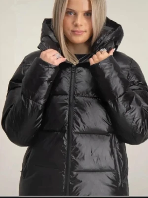 Svart pufferjacka med huva - Säljer en svart pufferjacka med glansigt material och stor huva. Jackan har dragkedja framtill och quiltade paneler som ger ett coolt och modernt uttryck. Perfekt för kalla vinterdagar och riktigt varm. Passar dig som gillar en stilren look.