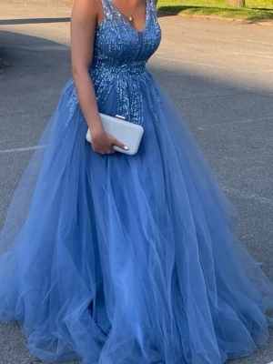 Balklänning blå - Elegant blå långklänning med v-ringning och glittriga paljettdetaljer upptill. Klänningen har en voluminös kjol i flera lager tyll och är ärmlös. Perfekt för bal eller speciella tillfällen.