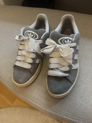Adidas Campus grå sneakers - Säljer ett par grå Adidas Campus sneakers med klassiska vita ränder och vit sula. Skorna är använda och har en tillagd tygbit vid hälen pga slitage, men annars superfina och otroligt sköna!