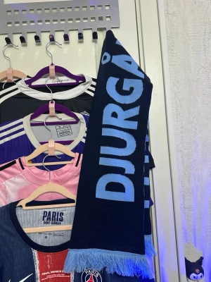 Blå Djurgården halsduk - Säljer en blå halsduk med texten 'DJURGÅRDEN' i ljusblått på ena sidan och blåa ränder på andra. Halsduken har ljusblå fransar i ändarna och är perfekt för att visa ditt stöd på matchen. Klassisk supporteraccessoar för Djurgården.