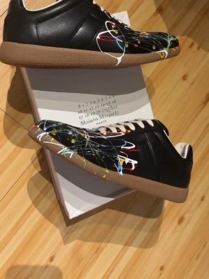 Maison Margiela sneakers med färgstänk - Maison Margiela sneakers i svart läder med unika färgstänk i blått, gult, vitt och rött. Skorna har beige gummisula och vita skosnören. Snygg och konstnärlig design som sticker ut. Perfekta för dig som vill ha något annorlunda och trendigt.