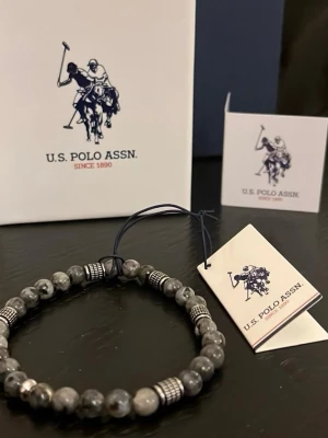 Herrarmband från U.S. Polo Assn. - Stilrent armband från U.S. Polo Assn. med gråmelerade pärlor och detaljer i silverfärgad metall. Elastisk tråd gör det enkelt att ta på och av. Perfekt accessoar för en trendig look.