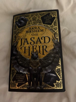 The Jasad Heir av Sara Hashem - Säljer fantasyromanen 'The Jasad Heir' av Sara Hashem. Boken har ett svart och guldigt omslag med detaljerad illustration av en bevingad varelse. Engelskspråkig, pocketutgåva. Perfekt för dig som gillar episk fantasy med magi, kungariken och förbjudna krafter.
