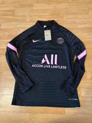 Svart PSG träningströja Nike - Svart långärmad träningströja från Nike för PSG med rosa detaljer och logga. Tröjan har halv dragkedja, klubbmärke på bröstet och texten 'Accor Live Limitless' framtill. Tillverkad i Dri-FIT-material som andas.