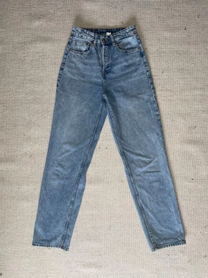 Blå mom jeans med hög midja - Klassiska blå mom jeans i 100% bomull med hög midja och raka ben. Jeansen har fem fickor, dragkedja och knapp framtill samt ljus tvätt för en retro känsla. Perfekta för en avslappnad och trendig look.